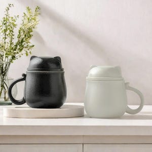 May include: Two ceramic mugs with cat-shaped lids. One mug is black and the other is white.
