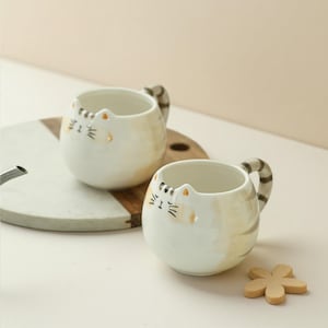 Peut inclure: Deux tasses en céramique blanche en forme de chat avec des marques brunes et orange. Les tasses ont un visage de chat avec des yeux noirs et des moustaches sur le devant. Les tasses sont posées sur une surface en bois.