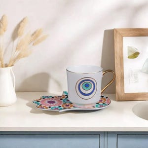 Puede incluir: Juego de taza y platillo de té de cerámica blanca. La taza presenta un asa dorada y un diseño de ojo azul, verde y dorado. El platillo tiene un estampado floral colorido. El juego está sobre una superficie blanca.
