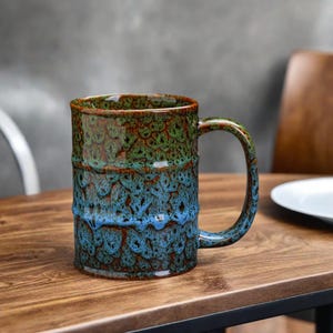 May include: A large, blue and green ceramic mug with a speckled glaze. The mug has a wide, rounded handle and a cylindrical shape.