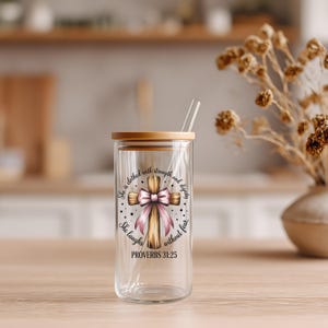 May include: A clear glass tumbler with a wooden lid and straw. The tumbler features a cross design with a pink bow and the text "She is clothed with strength and dignity. She laughs without fear. PROVERBS 31:25."
