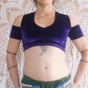 Peut inclure: Un haut court en velours violet avec des manches épaules nues et une longue jupe verte à volants. La jupe est assortie d'une ceinture décorative avec des ornements bleus, blancs et dorés, et des pompons noirs. Le fond est une tapisserie à motifs.
