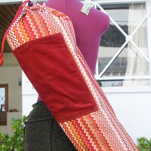 May include: A yoga mat bag with a red corduroy pocket and a chevron pattern in red, orange, and white. The bag is cylindrical and has a drawstring closure. The bag is displayed on a purple mannequin.