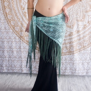 Puede incluir: Un top corto plateado de un solo hombro y una falda larga negra. Un pañuelo de cadera de encaje turquesa con flecos largos está envuelto alrededor de la cintura. Brazaletes dorados añaden un toque de elegancia. El fondo presenta un tapiz estampado.