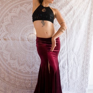May include: A woman wearing a black sequined halter top and a long, burgundy velvet skirt with a slit. She is standing in front of a white mandala tapestry.