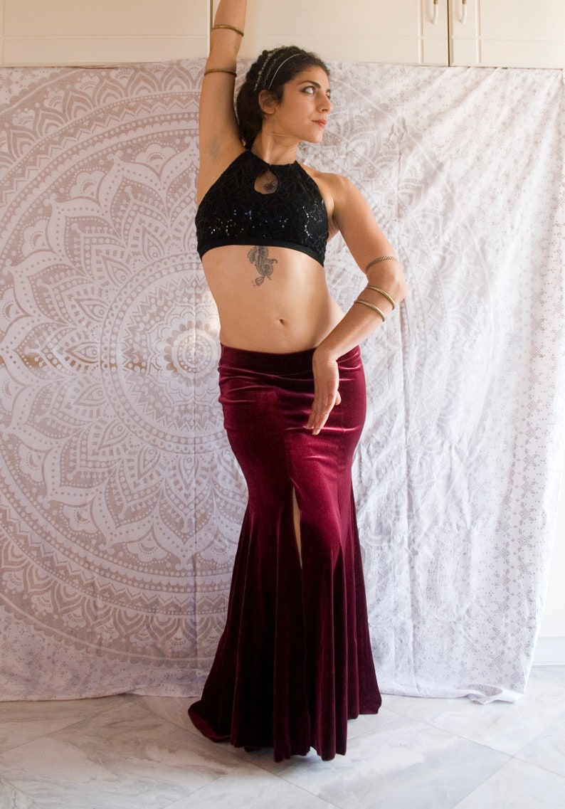 May include: A woman wearing a black sequined halter top and a long, burgundy velvet skirt with a slit. She is standing in front of a white mandala tapestry.