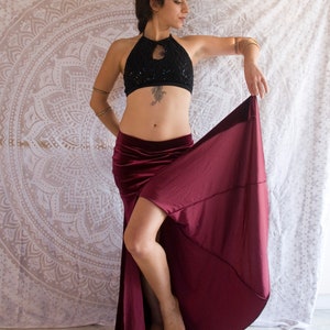 May include: A woman wearing a black sequined halter top and a burgundy velvet skirt with a high slit. She is posing with her skirt held up, revealing her legs.