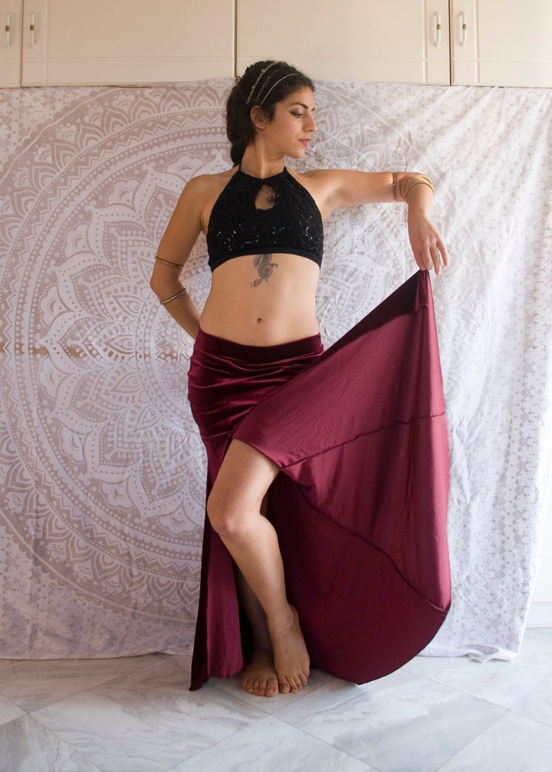 May include: A woman wearing a black sequined halter top and a burgundy velvet skirt with a high slit. She is posing with her skirt held up, revealing her legs.