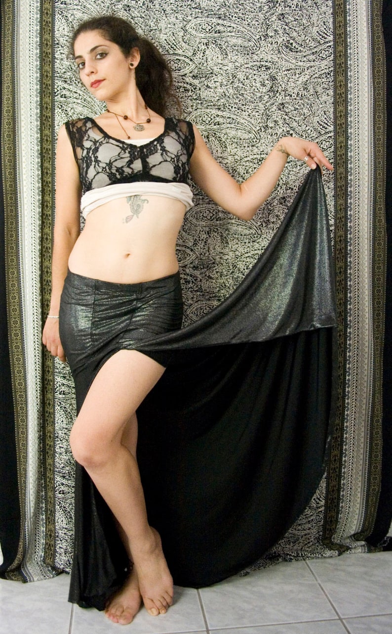 May include: A woman wearing a black lace top and a black skirt with a silver sheen. She is standing in front of a patterned background.