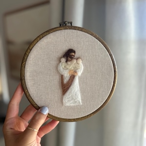 May include: A circular embroidery hoop containing a needle-felted textile art piece on light beige fabric. The design features a figure with dark brown hair wearing a white robe with a brown sash, holding two small infants wrapped in white.