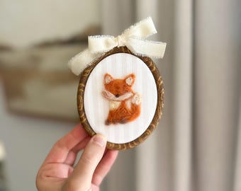 May include: Oval-shaped embroidery hoop featuring a detailed orange fox with a cream-colored bow. The fox is centered on a white fabric with vertical stripes, set within a wooden frame. A cream-colored ribbon is tied at the top.