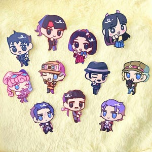 May include: A collection of colorful anime-style character stickers. Each sticker features a unique character design with detailed outfits and expressions. The stickers are arranged on a soft, yellow surface.