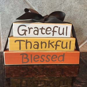 May include: Three wooden blocks stacked on top of each other, tied with a black ribbon. The blocks are painted in different colors: white, yellow, and orange. The words "Grateful", "Thankful", and "Blessed" are written in brown on the blocks.