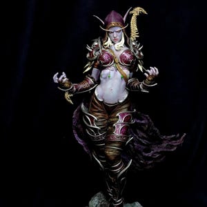 May include: A detailed figurine of a fantasy character with pale skin, wearing ornate armour in shades of burgundy, gold, and brown. The character has long, white hair, pointed ears, and a hooded head covering. The figure is posed against a black background.