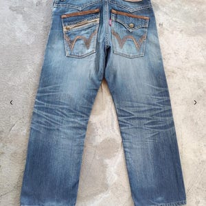 Peut inclure: Vue arri&egrave;re d'un jean en denim bleu avec des d&eacute;tails en cuir marron sur les poches et la ceinture. Le jean pr&eacute;sente un d&eacute;lavage et des coutures d&eacute;coratives sur les poches arri&egrave;re. Un patch en cuir rouge et marron est visible &agrave; la taille.