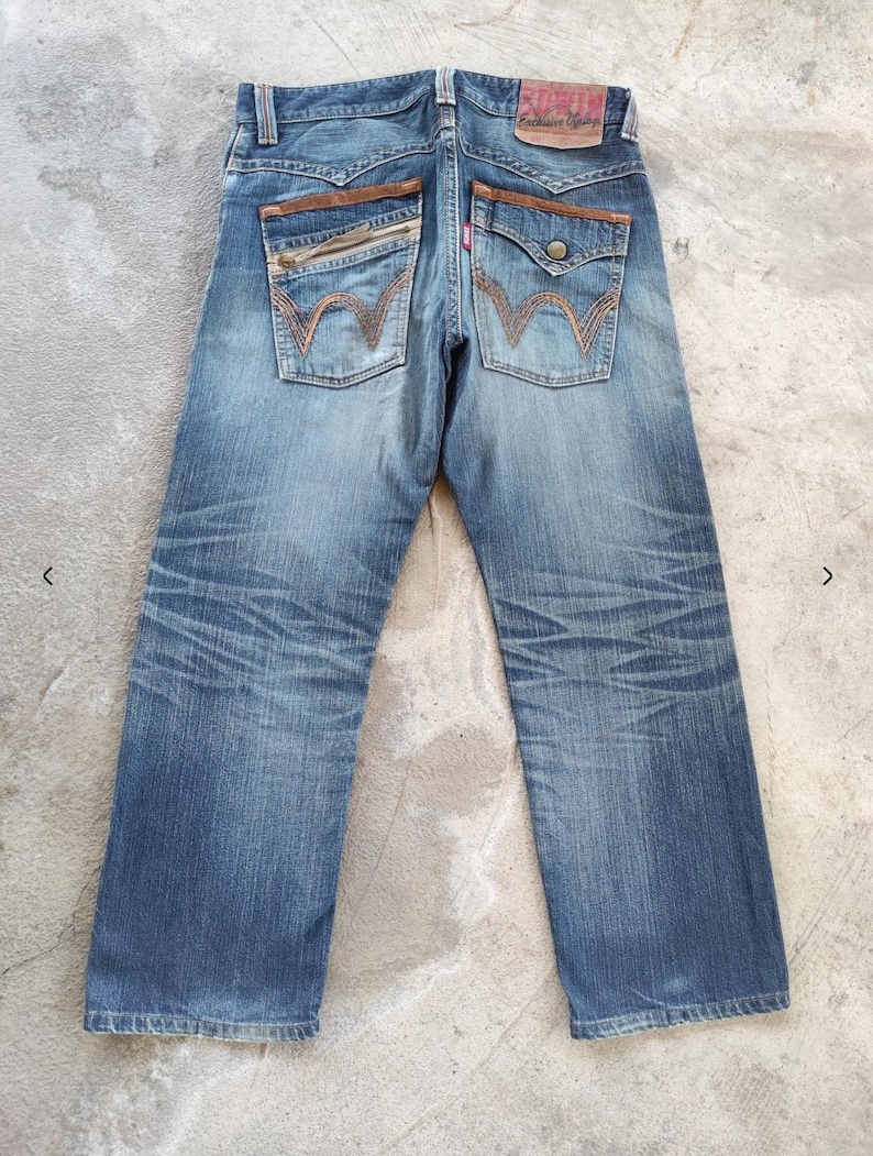 Peut inclure: Vue arri&egrave;re d'un jean en denim bleu avec des d&eacute;tails en cuir marron sur les poches et la ceinture. Le jean pr&eacute;sente un d&eacute;lavage et des coutures d&eacute;coratives sur les poches arri&egrave;re. Un patch en cuir rouge et marron est visible &agrave; la taille.