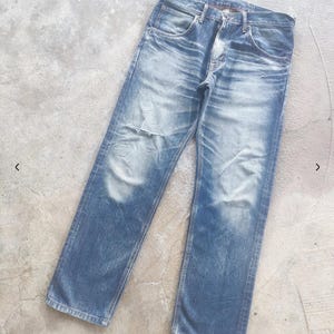 Puede incluir: Vaqueros de mezclilla azul descolorido con un aspecto desgastado. Los vaqueros tienen bragueta con botones, dise&ntilde;o cl&aacute;sico de cinco bolsillos y un parche desgastado en la rodilla izquierda. La mezclilla tiene un lavado claro con decoloraci&oacute;n y desgaste visibles.