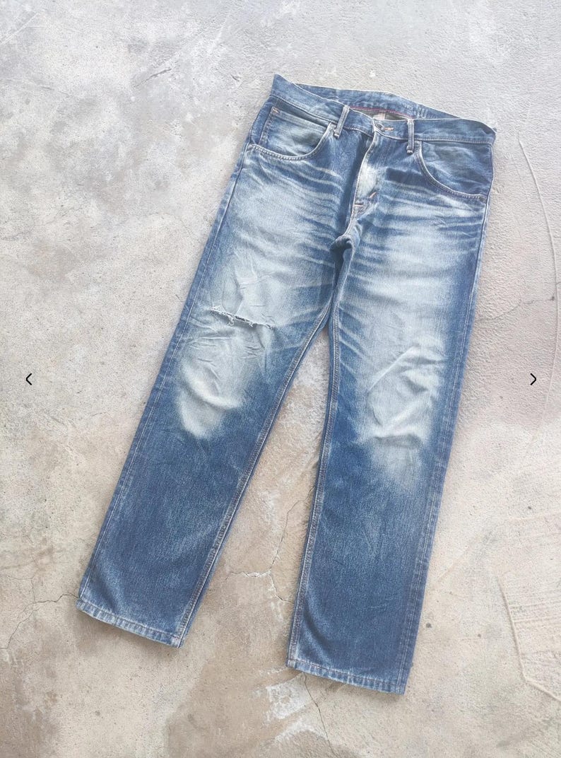 Puede incluir: Vaqueros de mezclilla azul descolorido con un aspecto desgastado. Los vaqueros tienen bragueta con botones, dise&ntilde;o cl&aacute;sico de cinco bolsillos y un parche desgastado en la rodilla izquierda. La mezclilla tiene un lavado claro con decoloraci&oacute;n y desgaste visibles.