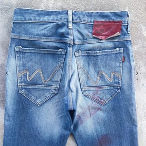 Puede incluir: Primer plano de la parte trasera de unos vaqueros azules. Los vaqueros tienen dos bolsillos traseros con un dise&ntilde;o de costura ondulada &uacute;nico. Un parche rojo es visible en la cintura. El denim tiene un aspecto descolorido y desgastado.