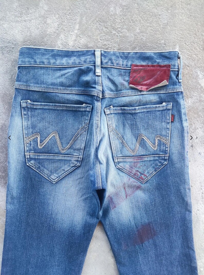 Puede incluir: Primer plano de la parte trasera de unos vaqueros azules. Los vaqueros tienen dos bolsillos traseros con un dise&ntilde;o de costura ondulada &uacute;nico. Un parche rojo es visible en la cintura. El denim tiene un aspecto descolorido y desgastado.