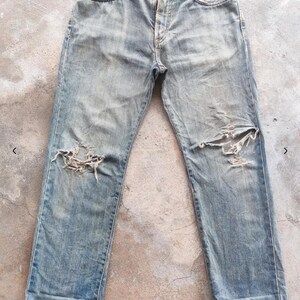 May include: Faded blue denim jeans with distressed detailing, including ripped knees. The jeans feature a classic five-pocket design and a button and zip closure. The fabric shows signs of wear.