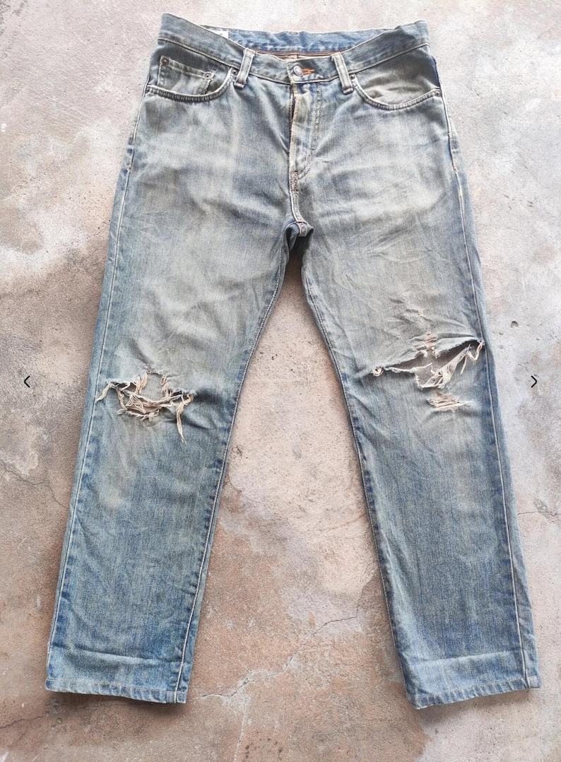 May include: Faded blue denim jeans with distressed detailing, including ripped knees. The jeans feature a classic five-pocket design and a button and zip closure. The fabric shows signs of wear.