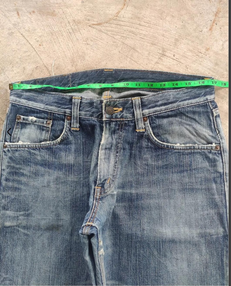 Puede incluir: Primer plano de unos vaqueros de mezclilla azules con cierre de bot&oacute;n y dise&ntilde;o cl&aacute;sico de cinco bolsillos. La cintura mide aproximadamente 40.6 cm de ancho. Los vaqueros muestran signos de desgaste y decoloraci&oacute;n, d&aacute;ndoles un aspecto vintage.