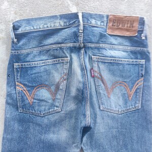 Puede incluir: Vaqueros de mezclilla azul descolorido con un parche de cuero marr&oacute;n que dice "EDWIN" en la parte trasera. Los bolsillos traseros tienen un dise&ntilde;o de costura naranja distintivo. Una peque&ntilde;a etiqueta roja es visible.
