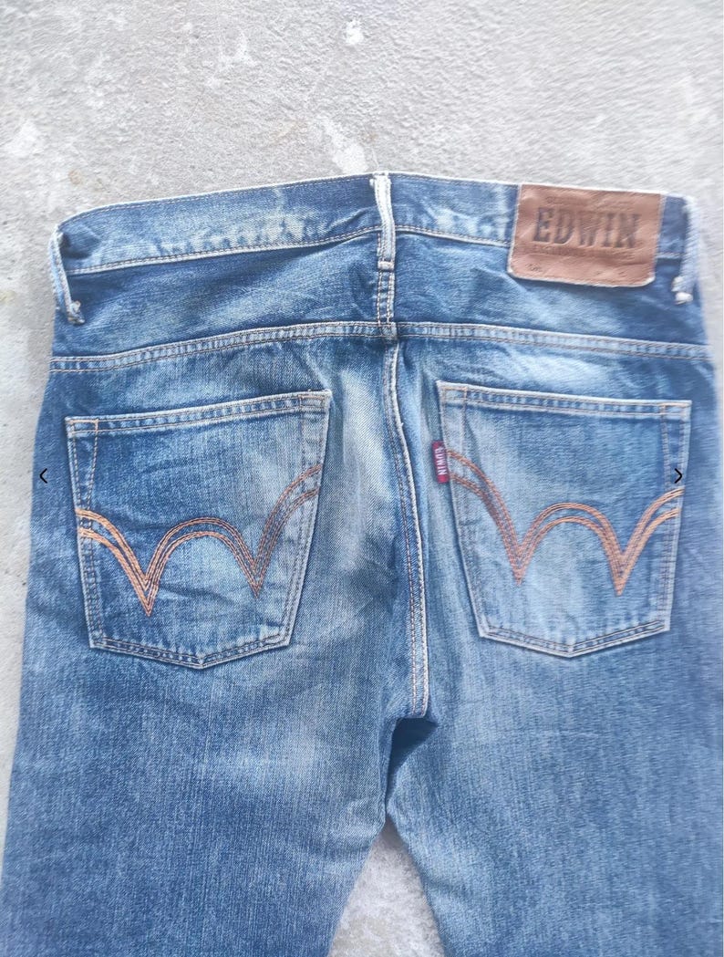 Puede incluir: Vaqueros de mezclilla azul descolorido con un parche de cuero marr&oacute;n que dice "EDWIN" en la parte trasera. Los bolsillos traseros tienen un dise&ntilde;o de costura naranja distintivo. Una peque&ntilde;a etiqueta roja es visible.