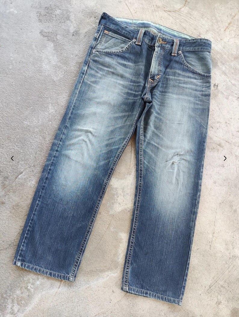 Peut inclure: Jean bleu d&eacute;lav&eacute; avec une conception classique &agrave; cinq poches. Le jean a une coupe droite et montre des signes d'usure, avec des zones plus claires sur les cuisses et les genoux. Le jean est pos&eacute; &agrave; plat sur une surface en b&eacute;ton.