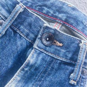 Puede incluir: Primer plano de unos vaqueros azules. Los vaqueros tienen cierre de bot&oacute;n, trabillas y costuras visibles. El denim tiene un aspecto descolorido y desgastado con una costura roja. El estilo sugiere ropa informal.