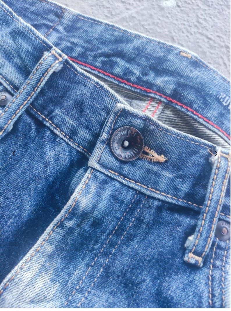 Puede incluir: Primer plano de unos vaqueros azules. Los vaqueros tienen cierre de bot&oacute;n, trabillas y costuras visibles. El denim tiene un aspecto descolorido y desgastado con una costura roja. El estilo sugiere ropa informal.