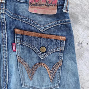 Peut inclure: Gros plan sur un jean en denim bleu avec un patch en cuir marron sur la poche arri&egrave;re. Le jean pr&eacute;sente une &eacute;tiquette rouge Edwin et une &eacute;tiquette en cuir marron avec l'inscription "Exclusive Vintage".