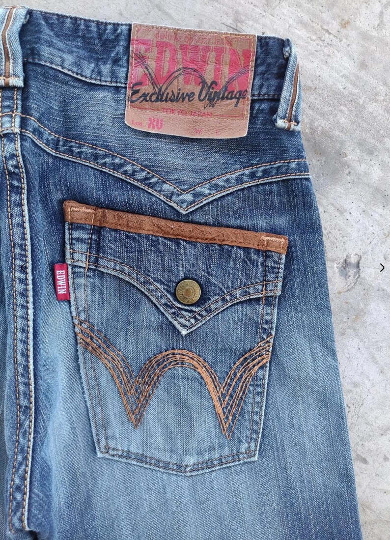 Peut inclure: Gros plan sur un jean en denim bleu avec un patch en cuir marron sur la poche arri&egrave;re. Le jean pr&eacute;sente une &eacute;tiquette rouge Edwin et une &eacute;tiquette en cuir marron avec l'inscription "Exclusive Vintage".