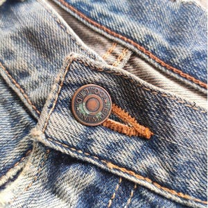 May include: Close-up of blue denim jeans with a button closure. The button is a tarnished copper colour and has the text "EDWIN CO." around the edge. The jeans have visible wear and tear, with orange stitching.