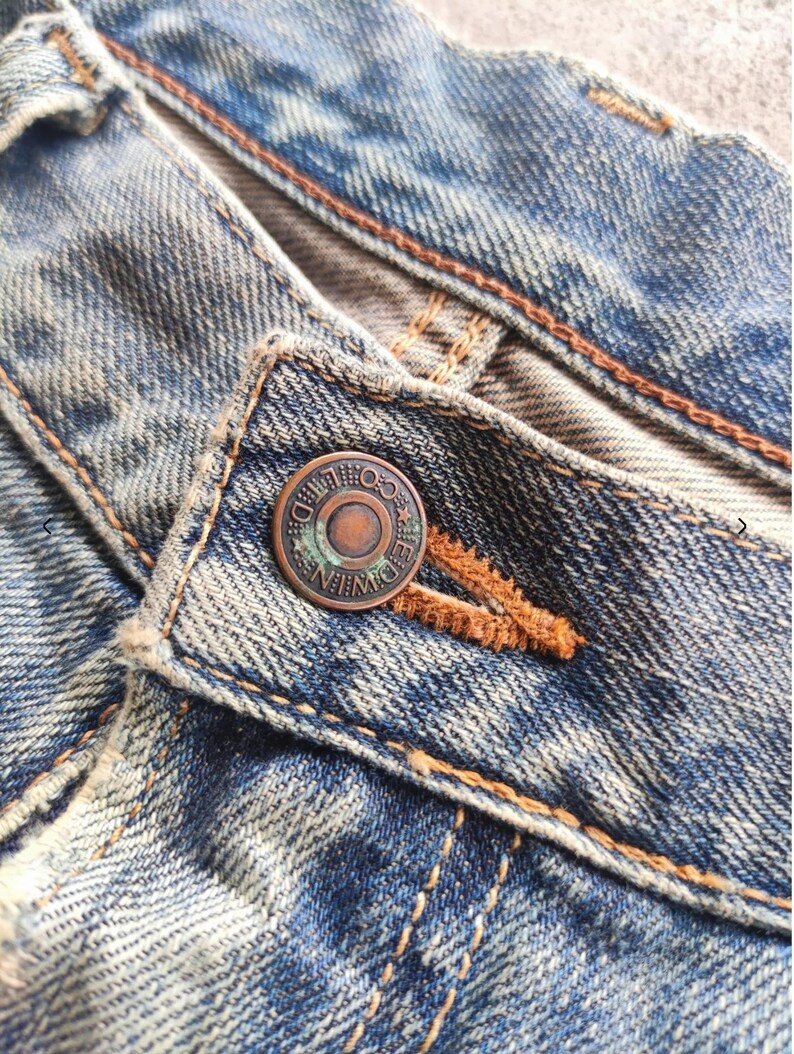 May include: Close-up of blue denim jeans with a button closure. The button is a tarnished copper colour and has the text "EDWIN CO." around the edge. The jeans have visible wear and tear, with orange stitching.