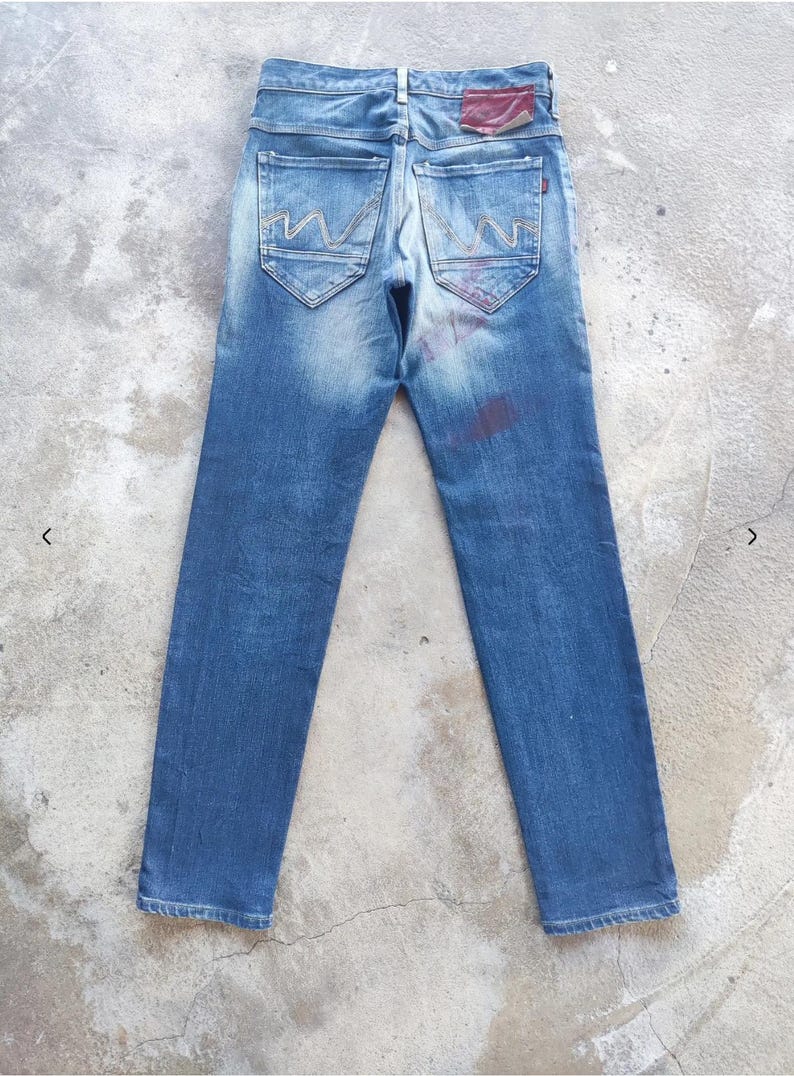 Puede incluir: Vaqueros azules con un lavado descolorido, costuras visibles y dise&ntilde;os decorativos en los bolsillos traseros. Presentan un parche rojo en la cintura trasera y un aspecto desgastado. Los vaqueros est&aacute;n colocados sobre una superficie de hormig&oacute;n.
