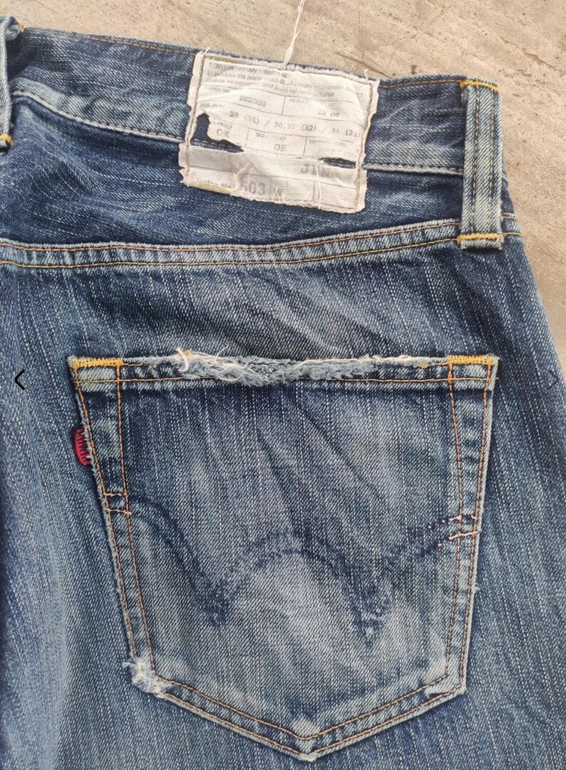 Puede incluir: Primer plano de la parte trasera de unos vaqueros azules. Los vaqueros tienen un bolsillo trasero con una etiqueta roja y un aspecto desgastado y envejecido. Una etiqueta blanca con texto es visible cerca de la cintura.