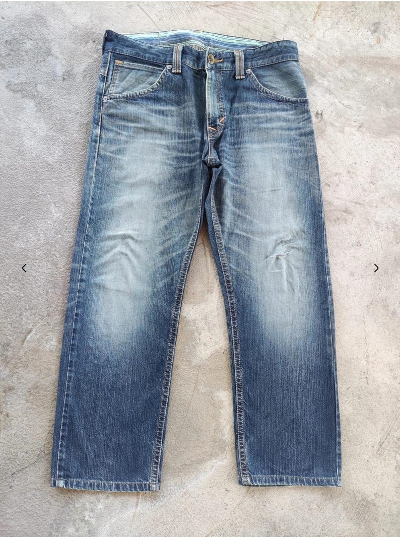 Peut inclure: Jean en denim bleu d&eacute;lav&eacute; avec une conception classique &agrave; cinq poches. Le jean a une coupe droite et pr&eacute;sente des signes d'usure, avec une d&eacute;coloration subtile et une zone us&eacute;e sur la jambe droite. La ceinture comporte des passants de ceinture et une fermeture boutonn&eacute;e.