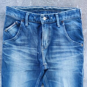 Puede incluir: Primer plano de unos vaqueros de mezclilla azules. Los vaqueros tienen un dise&ntilde;o cl&aacute;sico de cinco bolsillos con cierre de bot&oacute;n y cremallera. La mezclilla tiene un efecto de lavado descolorido, con zonas m&aacute;s claras en muslos y rodillas.