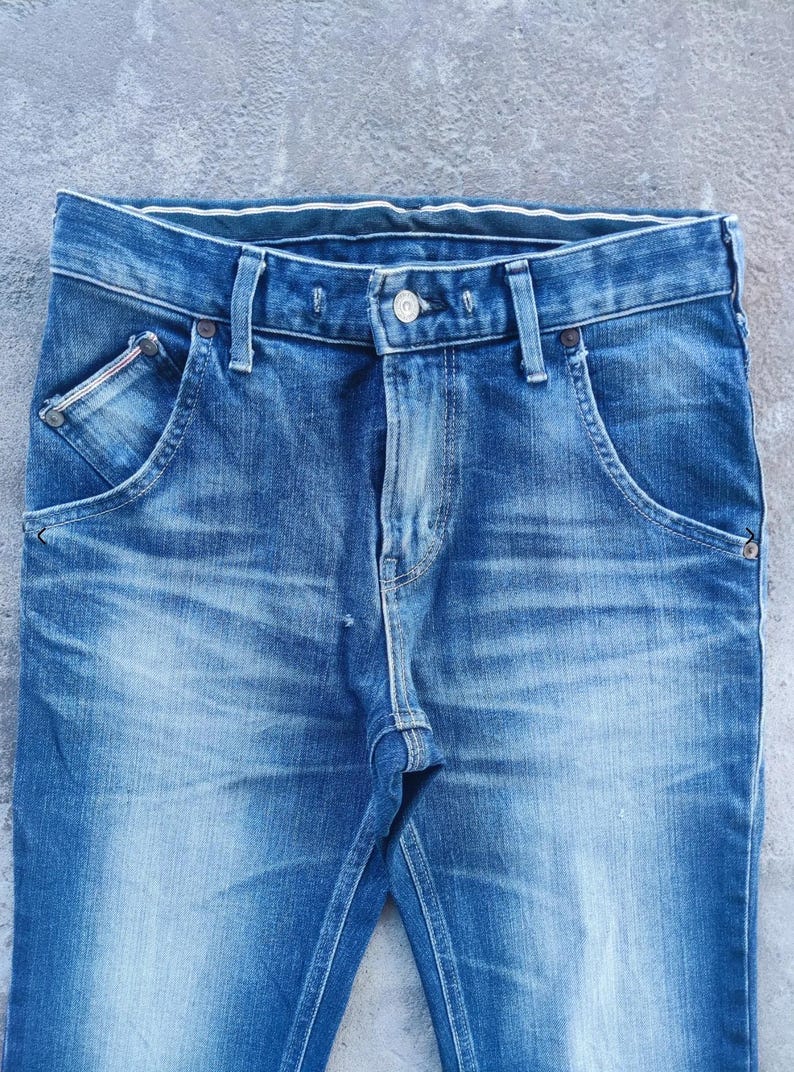 Puede incluir: Primer plano de unos vaqueros de mezclilla azules. Los vaqueros tienen un dise&ntilde;o cl&aacute;sico de cinco bolsillos con cierre de bot&oacute;n y cremallera. La mezclilla tiene un efecto de lavado descolorido, con zonas m&aacute;s claras en muslos y rodillas.