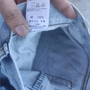 May include: Close-up of a light blue denim garment with a white tag. The tag has Japanese text and symbols, including washing instructions. The fabric is 100% cotton. The tag also indicates the use of cowhide leather.