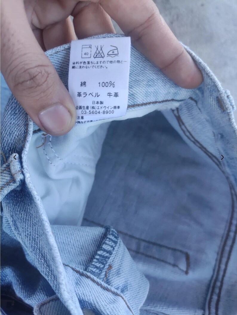 May include: Close-up of a light blue denim garment with a white tag. The tag has Japanese text and symbols, including washing instructions. The fabric is 100% cotton. The tag also indicates the use of cowhide leather.