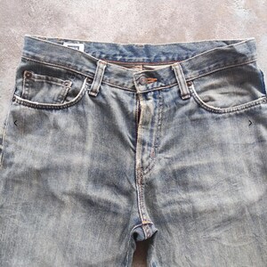 May include: Close-up of faded blue denim jeans. The jeans have a button closure, belt loops, and front and back pockets. The denim has a worn, distressed appearance.