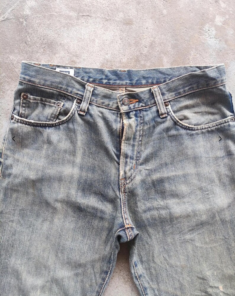 May include: Close-up of faded blue denim jeans. The jeans have a button closure, belt loops, and front and back pockets. The denim has a worn, distressed appearance.