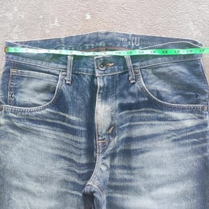 Puede incluir: Primer plano de unos vaqueros de mezclilla azules con cierre de bot&oacute;n y bolsillos. La cintura mide aproximadamente 40.6 cm de ancho. Los vaqueros tienen un lavado descolorido y la marca "EDWIN" es visible.