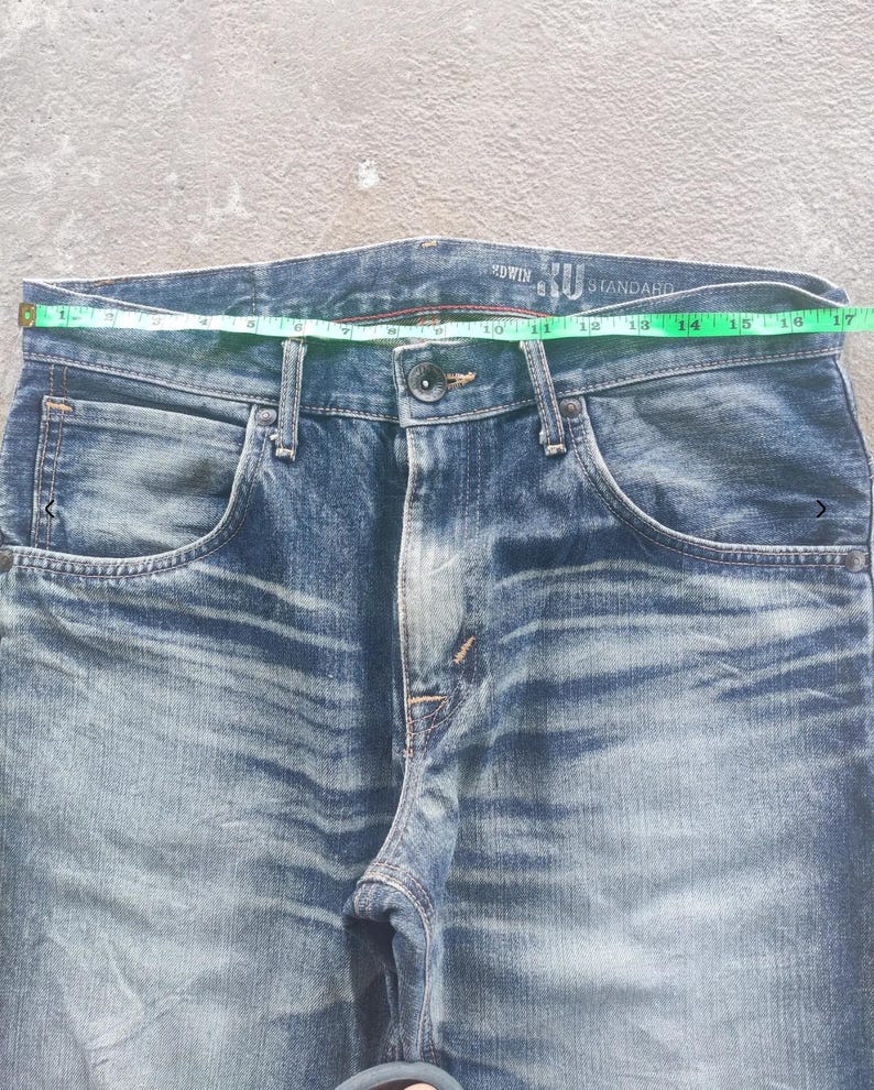 Puede incluir: Primer plano de unos vaqueros de mezclilla azules con cierre de bot&oacute;n y bolsillos. La cintura mide aproximadamente 40.6 cm de ancho. Los vaqueros tienen un lavado descolorido y la marca "EDWIN" es visible.
