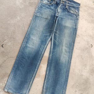 Puede incluir: Vaqueros de mezclilla azul descolorido con un dise&ntilde;o cl&aacute;sico de cinco bolsillos. Los vaqueros tienen un corte de pierna recta y muestran signos de desgaste, con &aacute;reas m&aacute;s claras y una decoloraci&oacute;n sutil. Los vaqueros est&aacute;n planos sobre una superficie de hormig&oacute;n.