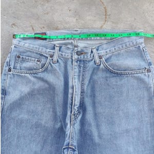 May include: Light blue denim jeans with a straight leg cut. A green measuring tape shows the waist size. The jeans have classic five-pocket styling with a button and zipper closure. The jeans are made of a light wash denim.