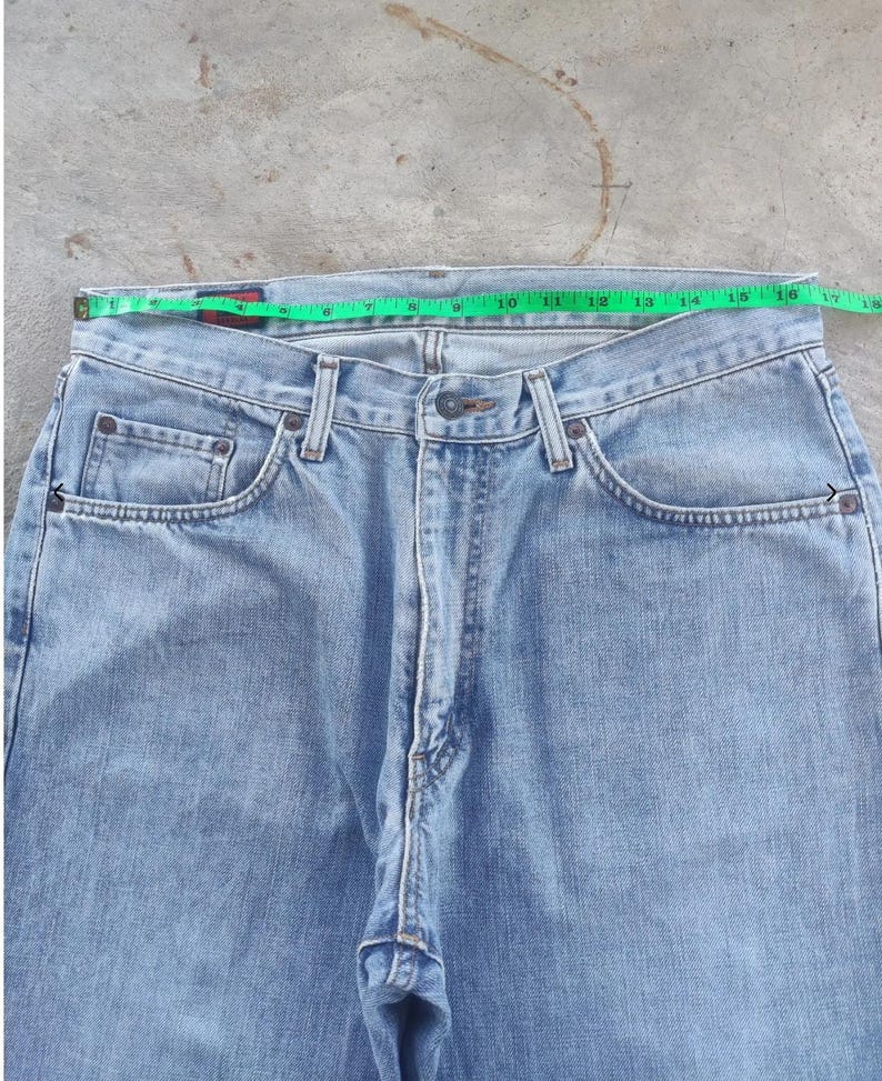 May include: Light blue denim jeans with a straight leg cut. A green measuring tape shows the waist size. The jeans have classic five-pocket styling with a button and zipper closure. The jeans are made of a light wash denim.